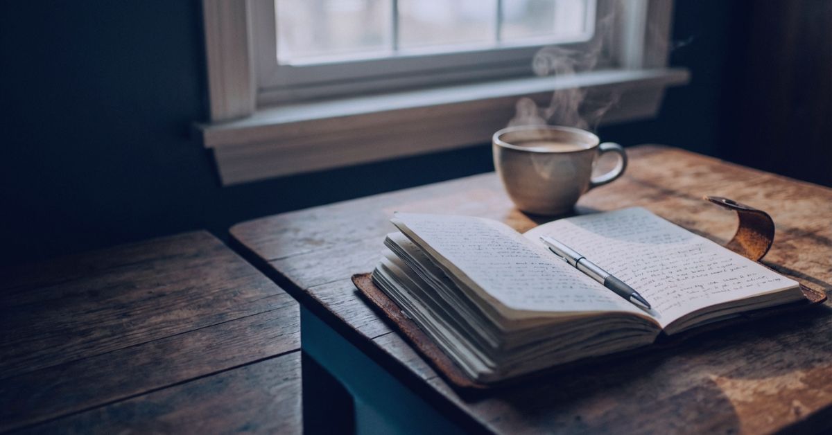 An open journal and pen beside a steaming cup by a window